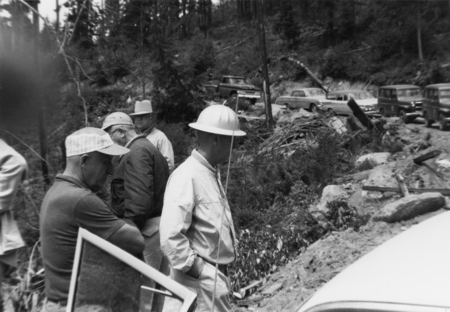 Several men wearing hats and jackets stand near a pile of debris in a forested area. There are cars parked on a dirt road in the background, and trees surround the scene.