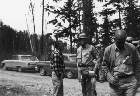 A group of men stand on a dirt road in a wooded area with parked cars nearby. One man, wearing a checkered shirt, appears to be speaking to another man holding a megaphone. Another man walks by holding a can. Tall trees surround the scene.