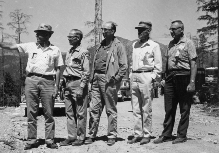 Five men stand on a dirt road, surrounded by trees and parked cars. They are wearing casual clothing, including hats, and appear to be engaged in a discussion, with one man pointing into the distance.