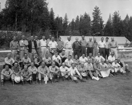 A group of men posed in two rows on a grassy area, with trees and a building in the background. Some are sitting on the ground while others stand behind them. They are dressed in casual shirts and trousers, and several hold hats or rolled papers.