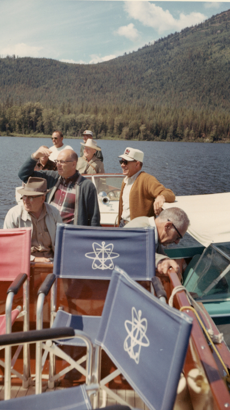 A group of men are standing on a boat surrounded by water with forested hills in the background. Some are wearing hats and sunglasses, and a few are seated on chairs with scientific symbols printed on them. The scene appears relaxed and casual.