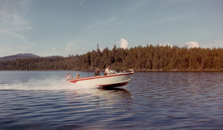 A motorboat with three people aboard is speeding across a lake. One person is driving, while two others sit in the boat. Trees line the opposite shore of the lake. An American flag is visible on the boat.