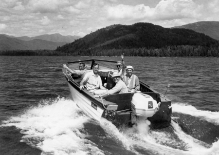 A group of five men is riding in a small boat on a lake. They are smiling and waving as the boat moves through the water. Behind them, there is a scenic view of hills and trees.