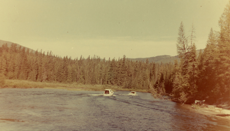 Two small boats are moving along a river, surrounded by a dense forest of tall trees with a mountain in the background.