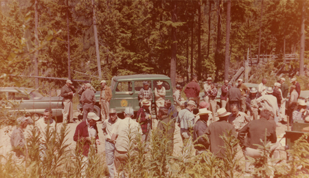A large group of people gathered outdoors in a forested area with tall trees. Several individuals are engaged in conversation near parked vehicles. Some people are wearing hats and casual clothing, and greenery is visible in the foreground.