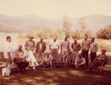 A group of men gathered outdoors on a grassy area with trees and mountains in the background. Some are sitting on chairs, while others are standing. They appear to be engaged in conversation or enjoying leisure time together.