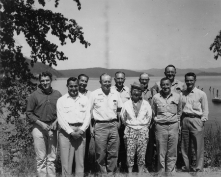 A group of nine people standing together near a body of water, with trees on either side and distant mountains in the background.