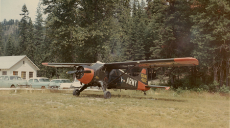 An airplane with markings on its side is parked on a grassy field. Behind it, there are several vintage cars and a building with a light-colored exterior. The area is surrounded by tall trees. National Guard plane at Cavanaugh Bay with text reading: "IDANG 41682" and "ARMY"