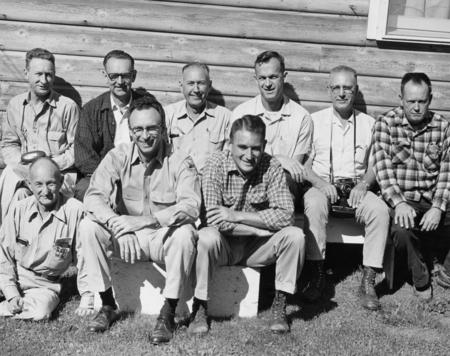 Nine men sitting and kneeling in two rows in front of a wooden building. Two men in the front row are seated on a bench, and one has a camera resting on his knee. Most are wearing shirts with collars and a few have on glasses. The setting is outdoors on grass.