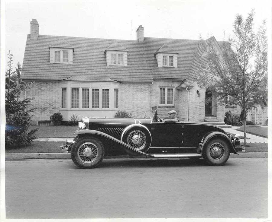 Photograph of view of Frank B. Robinson sitting in the driver's side of his Duesenberg convertible coupe at the curb in front of his house.