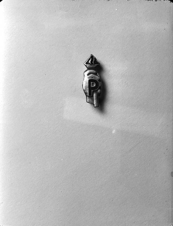 Negative photograph of a pin worn by members of Psychiana. Pin includes a small hand holding a lit torch with the capital letter 'P' prominently in front.