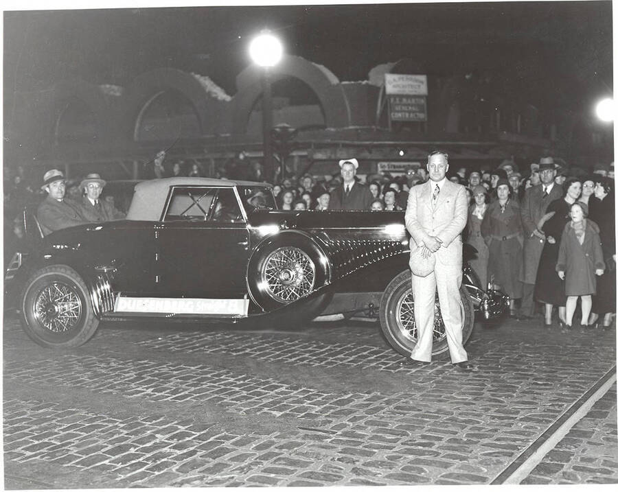 Photograph of Frank B. Robinson standing by the hood of his Duesenberg Convertible Coupe in a public square or street with many people standing behind him. Along the running board, a sign reads 'Duesenberg Straight 8.'
