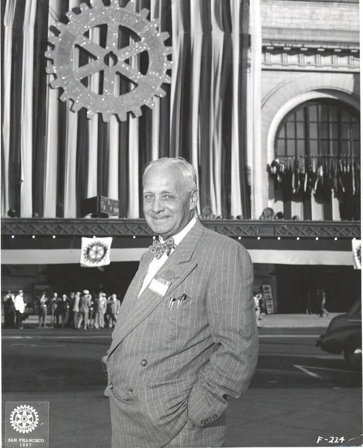 Photograph of older Frank B. Robinson stands beside a public street in a pin-striped suit and bowtie with his hands in his pockets.