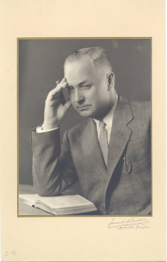 Portrait of middle-aged Frank B. Robinsons wearing a gray suit, not facing the camera and leaning over a book with a pen in the same hand he uses to rub his brow.