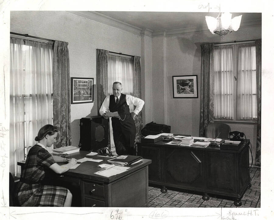 Photograph of Frank B. Robinson leaning against a bookshelf and dictating a document to a female Psychiana staff member writing on a piece of paper. Includes penciled dimensions in white margin.