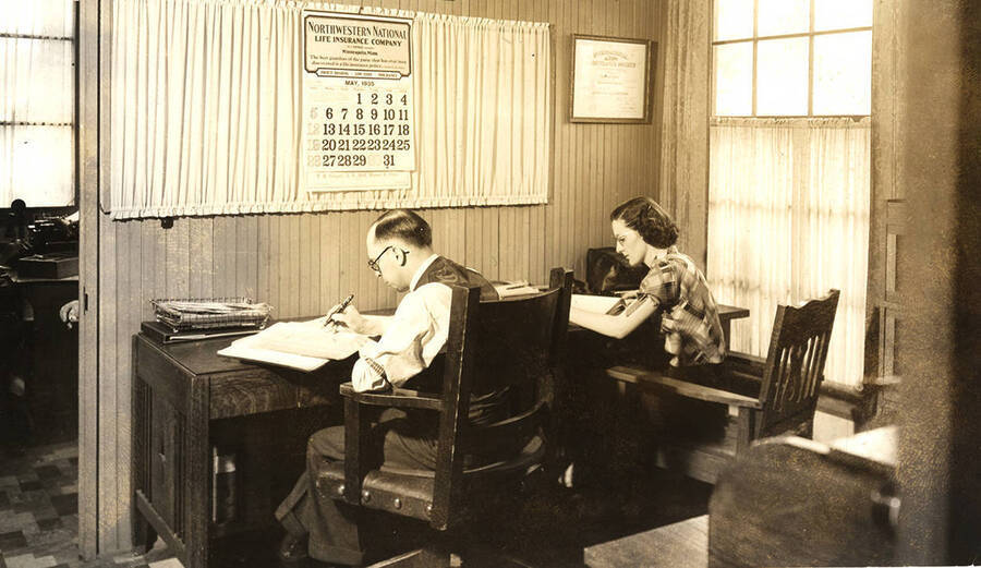 Photograph depicts an unidentified man and woman sitting over a desk and writing records in ledgers or some sort of record books. The calendar over the desk marks the month of May in year 1938.