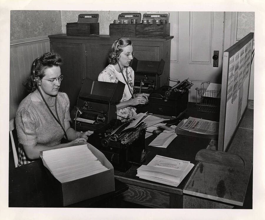 Photograph depicts two women sitting next to each other at a desk and using Dictaphones and typewriters to transcribe documents.