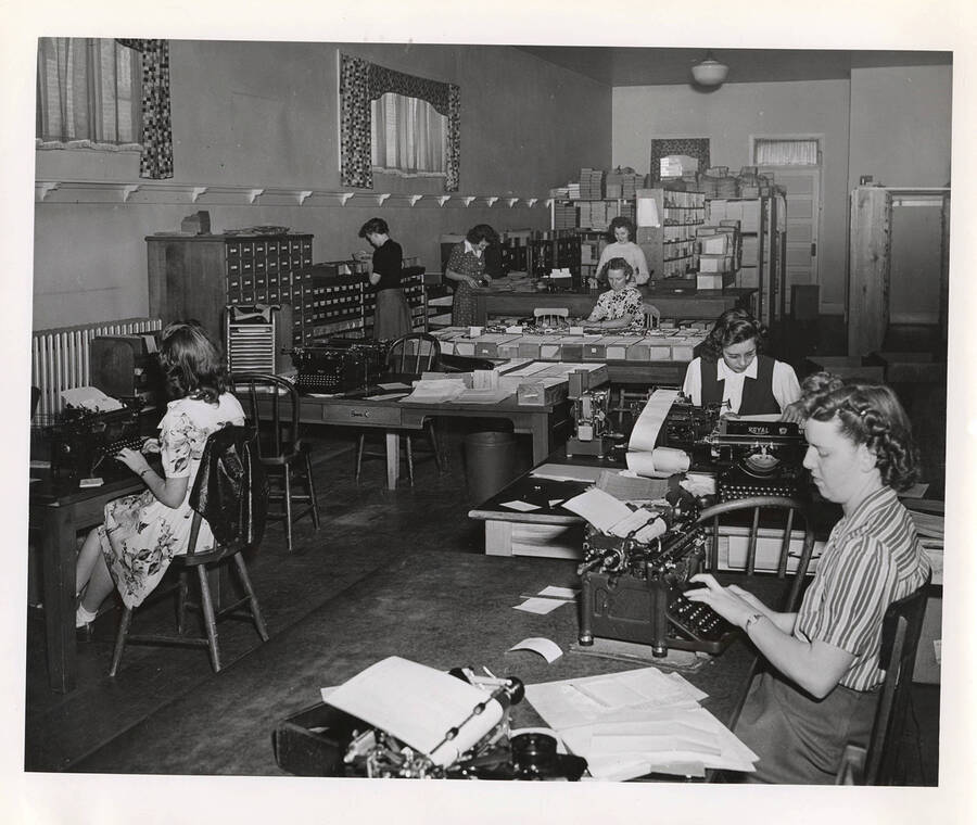 Photograph depicts the bookkeeping department in the Psychiana Branch Mailing Department and several female staff completing various tasks of typing, sorting mail, calculating and keeping records.