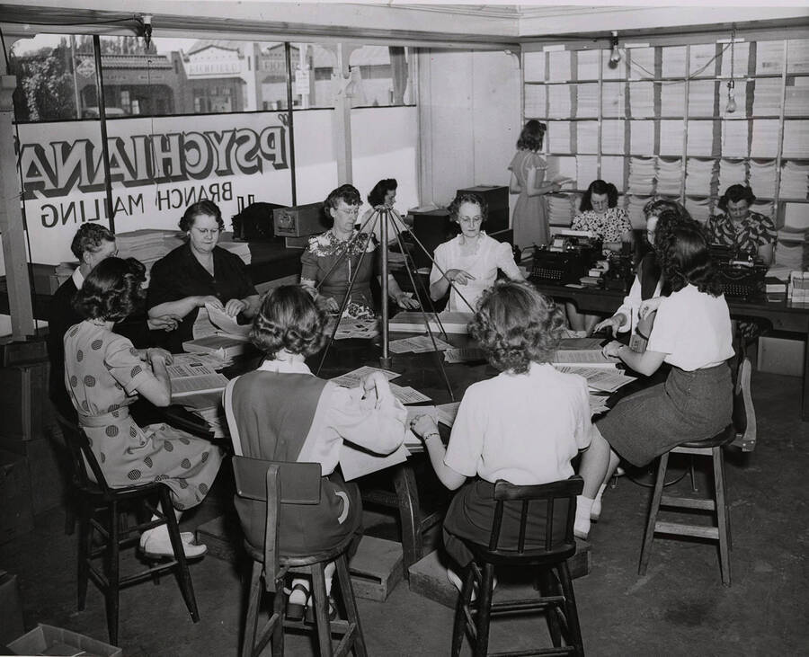 Photograph depicts several female Psychiana staff sitting around a revolving 'rotary table,' operated by electric motor and used to assemble Frank B. Robinsons Lessons and follow-up literature before mailing.