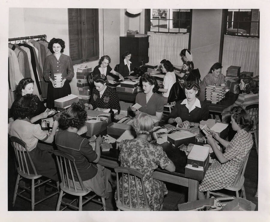 Photograph depicts a room full of female Psychiana staff sitting at long tables and working together to sort and compile material for mailing.