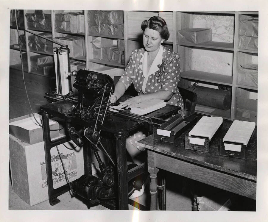 Photograph depicts a female Psychiana staff member sitting at a cutting or labeling machine and using it to prepare several envelopes for mailing.