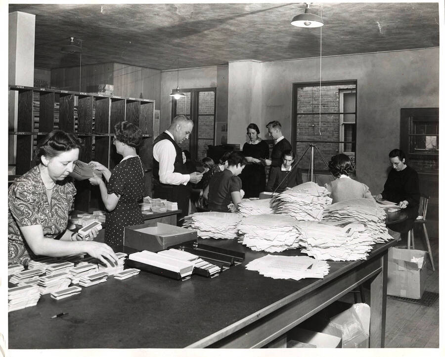 Photograph depicts Frank B. Robinson standing in the center of the mail room reading a piece of paper while Psychiana staff work around him sorting mail and compiling literature at a revolving rotary table.