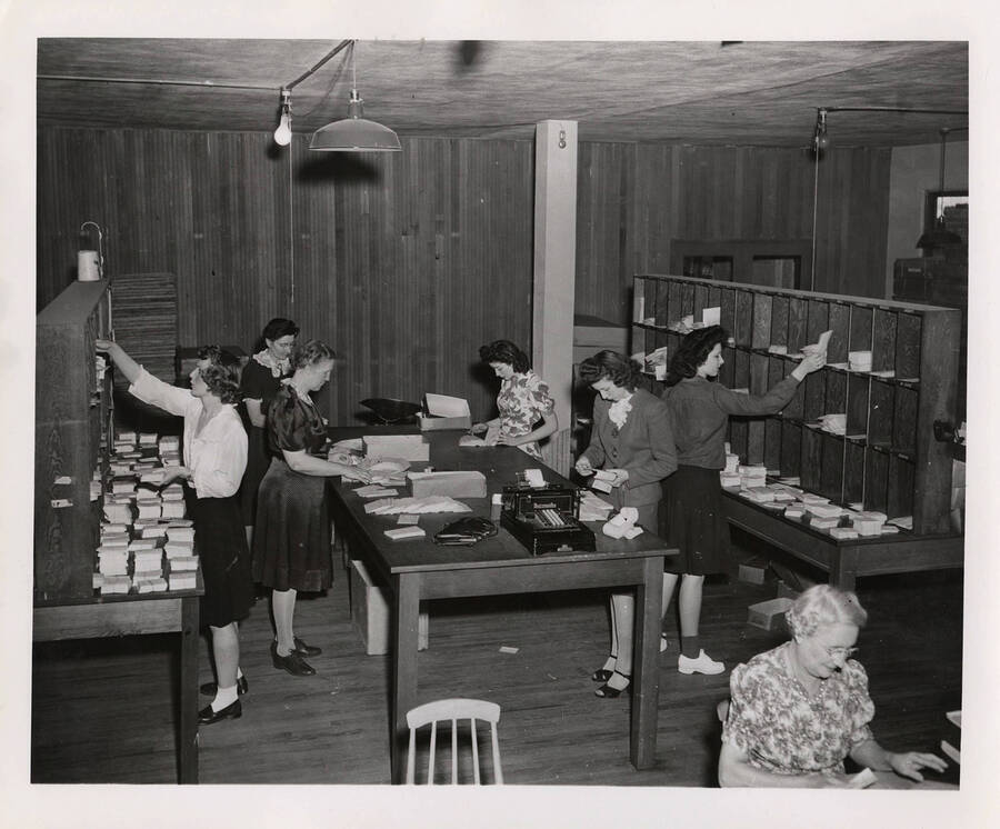 Photograph from a higher vantage depicts the mail room and several female Psychiana staff sorting letters at a central table and depositing envelopes into a lattice sorting shelf.