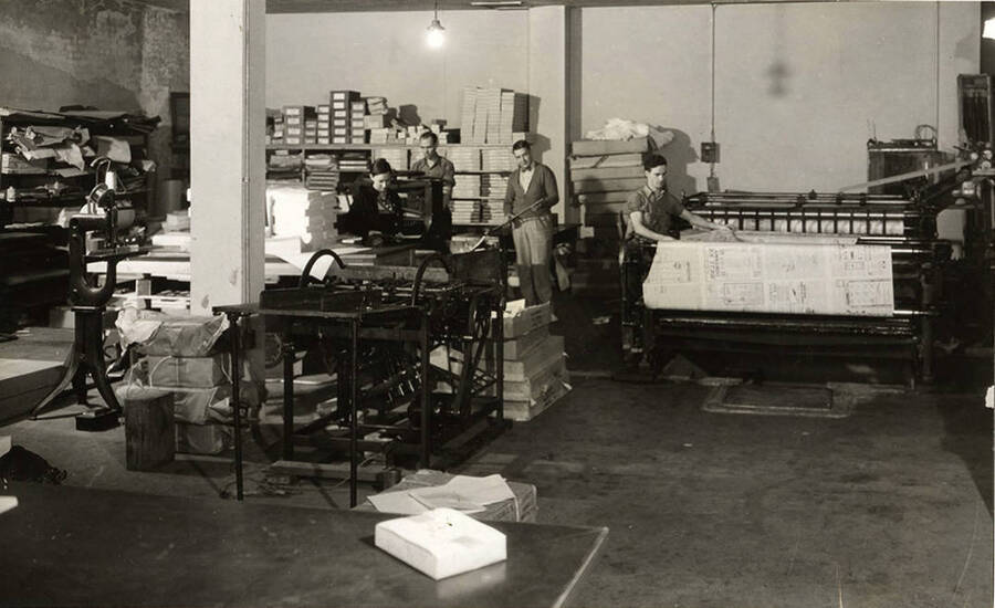Photograph depicts a large warehouse room in the Newspaper Plant with several workers sorting papers and working at a large industrial printing press.