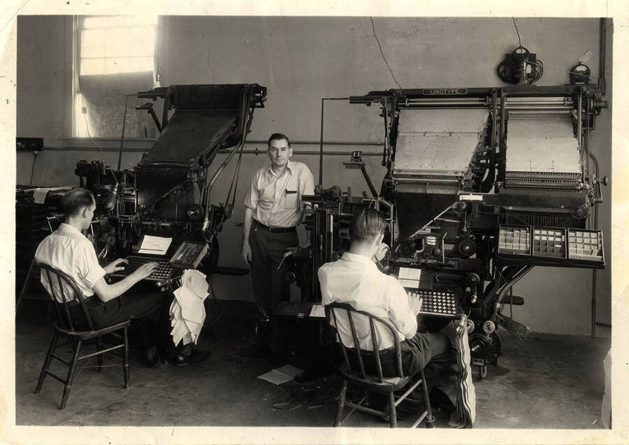 Photograph depicts W.J. Marinean standing between Howard Wallace and T.J. Grief, who sit at two linograph machines and work.