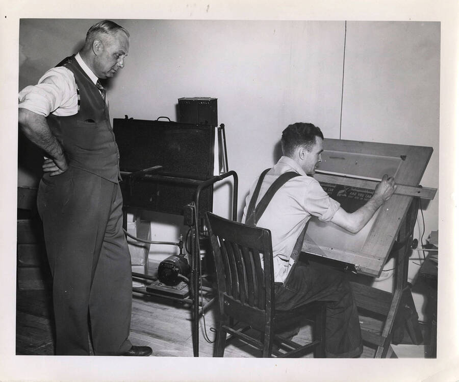 Photograph depicts Frank B. Robinson standing behind an unidentified male staff member who edits a lithograph plate at a drafting table.