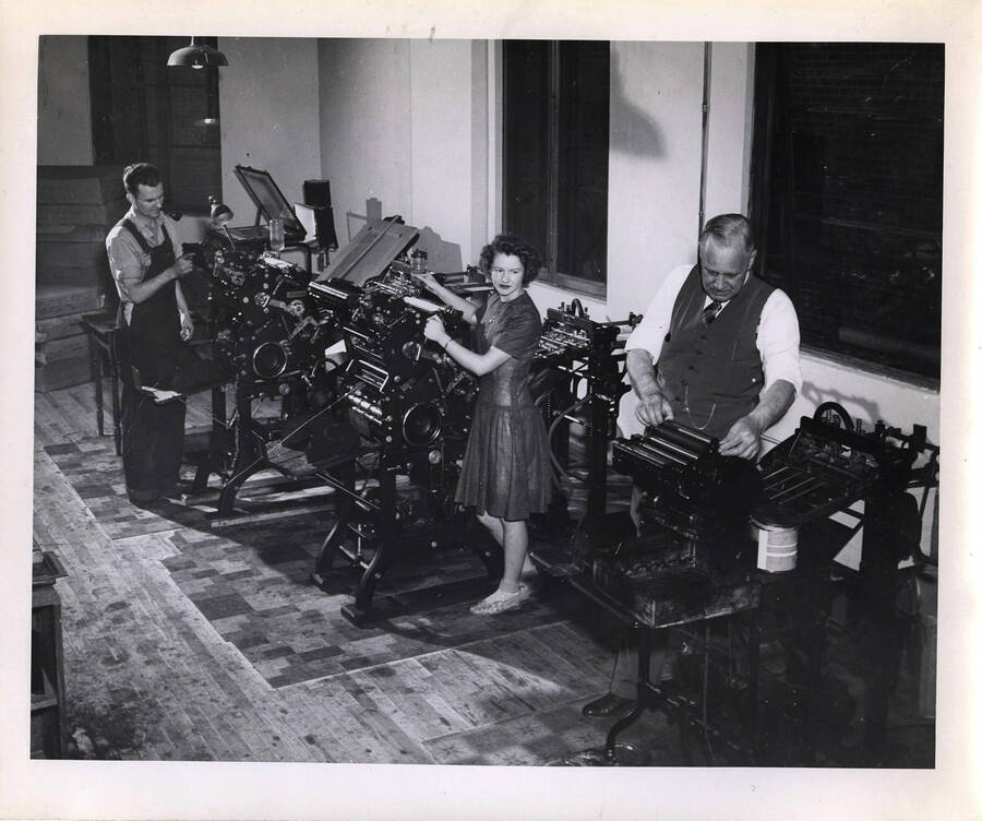 Photograph depicts Frank B. Robinson with his sleeves rolled up and an unidentified male and female staff member working at row of small printing presses.
