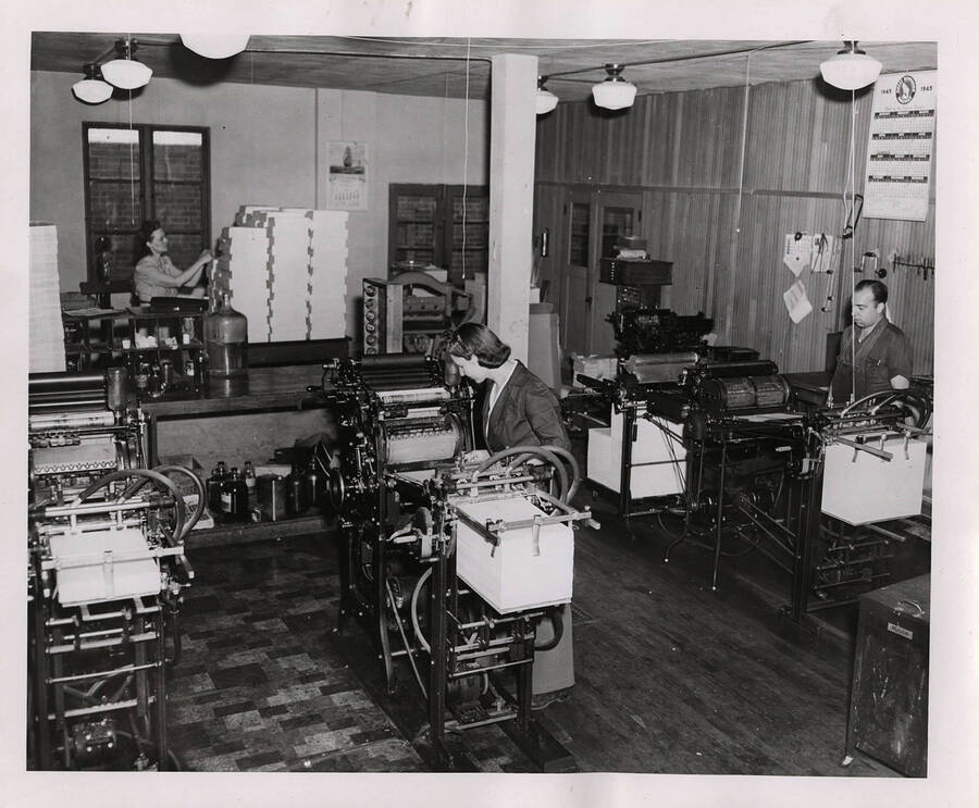 Photograph depicts several unidentified Psychiana employees working at several different machines, including small side by side printing presses, and sorting materials in the back of the room.