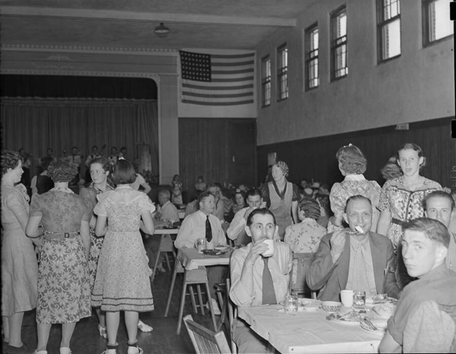 Photograph depicts dinner time inside the Uniontown Building during the Uniontown stock show, with several unidentified men and women sitting at tables and standing with beverages and plates.