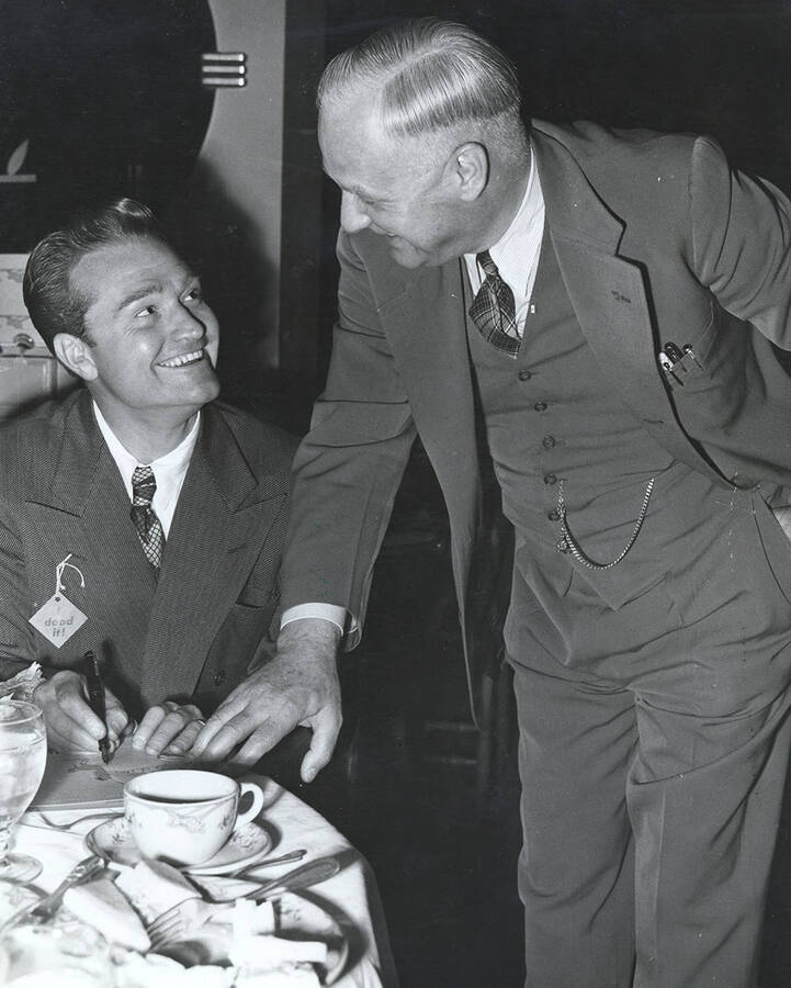 Photograph depicts Frank. B. Robinson standing over a dinner table and leaning down to talk to Red Skelton, who sits with a pen signing a piece of paper.