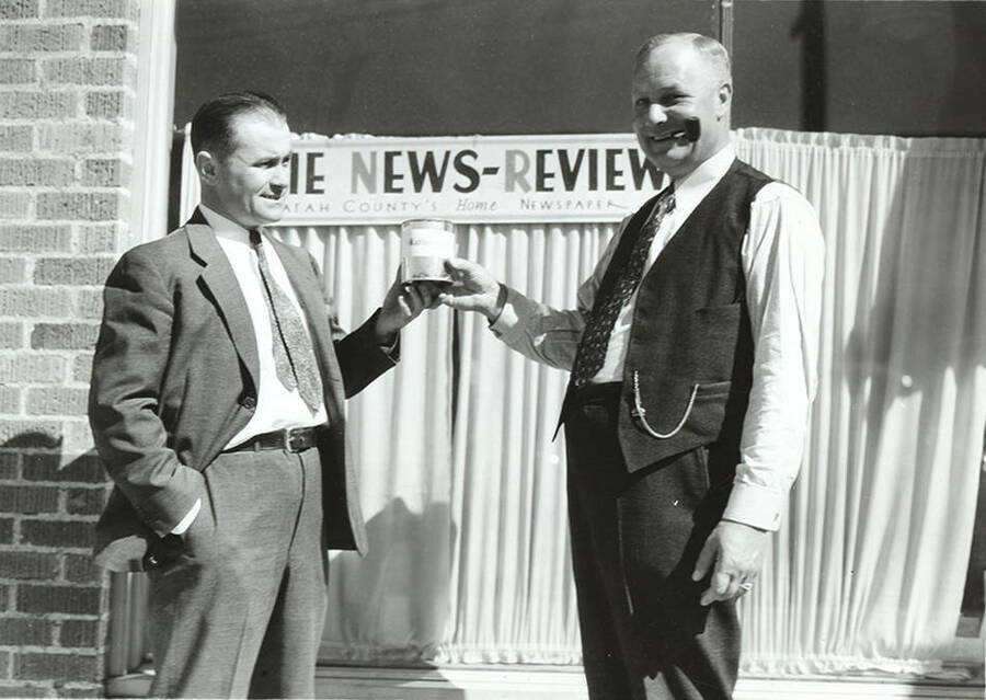 Photograph depicts Frank B. Robinson handing Mr. Henry Banks a can of 'rattlesnake meat' out front of the building of the publication, The News Review.