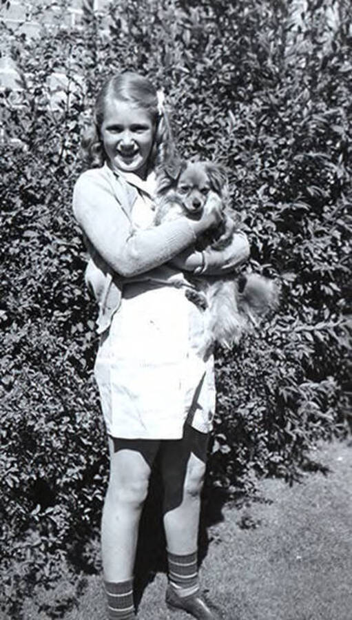 Photograph depicts Florence Robinson, age 11, standing outside in front of bushes and holding a small dog.