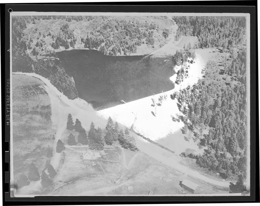 Negative photograph from overhead view of a lake either after or during its development. Photograph includes a storage structure and several dirt roads running around and into the lake.