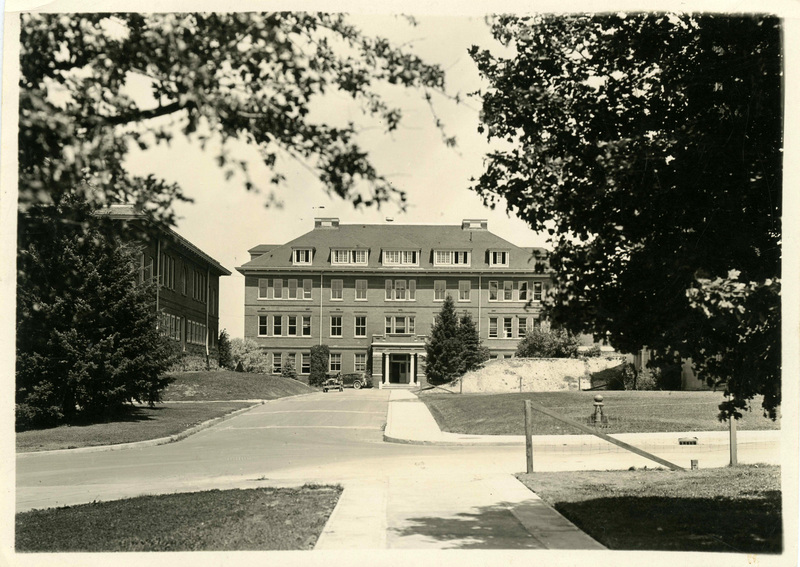 Looking north at Morrill Hall (center) from edge of Admin Lawn, College of Art and Architecture (formerly Metallurgical Building, left). Streets and sidewalk are paved, a wooden and wire fence is at the right edge of Admin Lawn, two old cars are near Morril Hall.