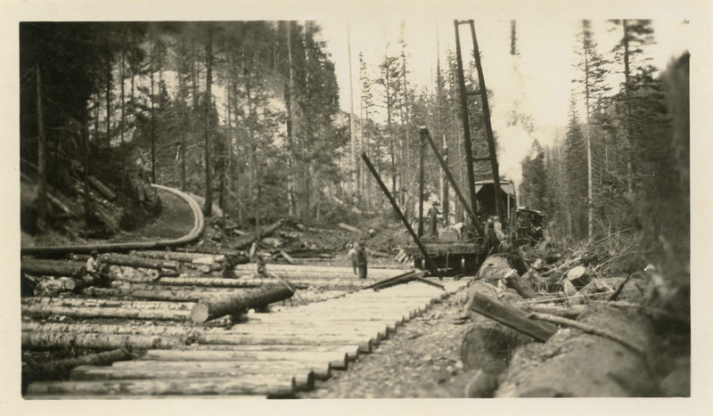Workers laying rails on a spur line in Burnt Cabin Creek, Coeur d'Alene National Forest. Cut logs lined up in front of a crane workers are using to move more logs in place. Forested hillside in background.