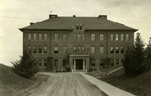 Looking north at Morrill Hall from a dirt road next to a paved sidewalk with grass covered hills  and trees on either side. Two old cars are parked in front of the building.