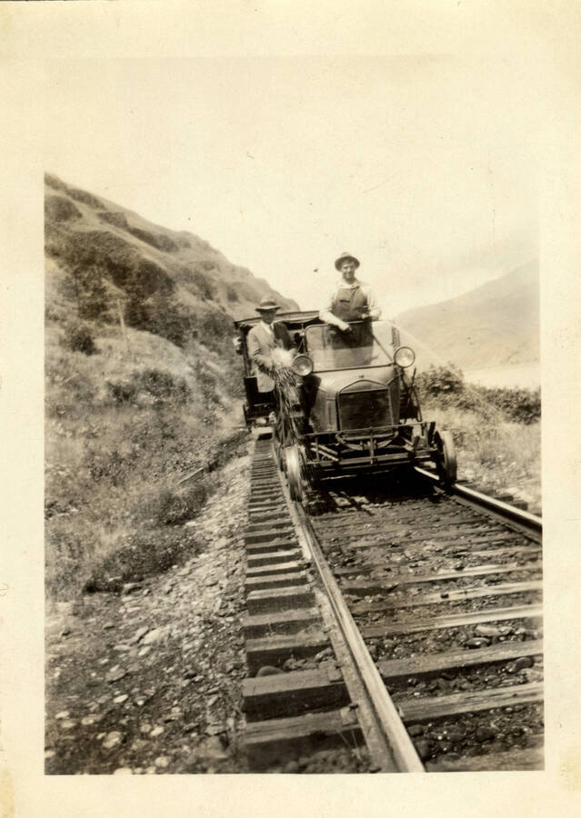 A photograph of a man driving a passenger car. The photo is taken headon, showing the car approaching. Pictured on the car are two men, one dressed in a professional suit, another in clothing that appears to suit that of a railroad worker.