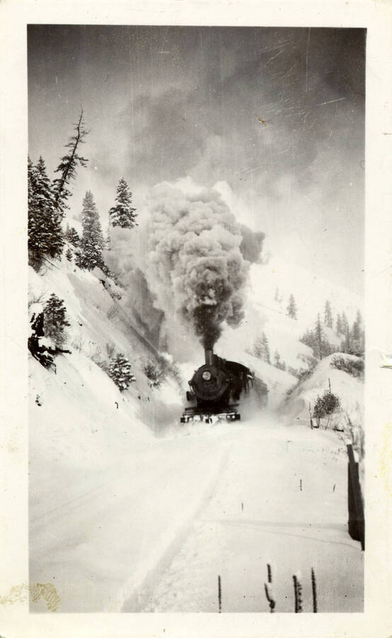 A photograph of a moving train engine huffing through the snow.