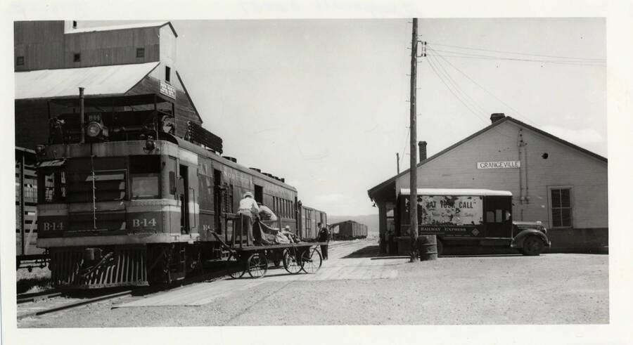 Northern Pacific B-14 railway post office locomotive pictured with a railway express truck being loaded with mail in Grangeville, Idaho. Two postal workers loading the locomotive from a rail cart.
