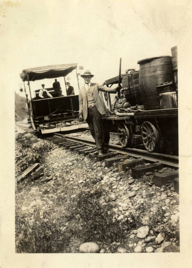A man holds a gun while standing next to a passenger car full of cargo and materials. Behind him is a construction car with three men inside.