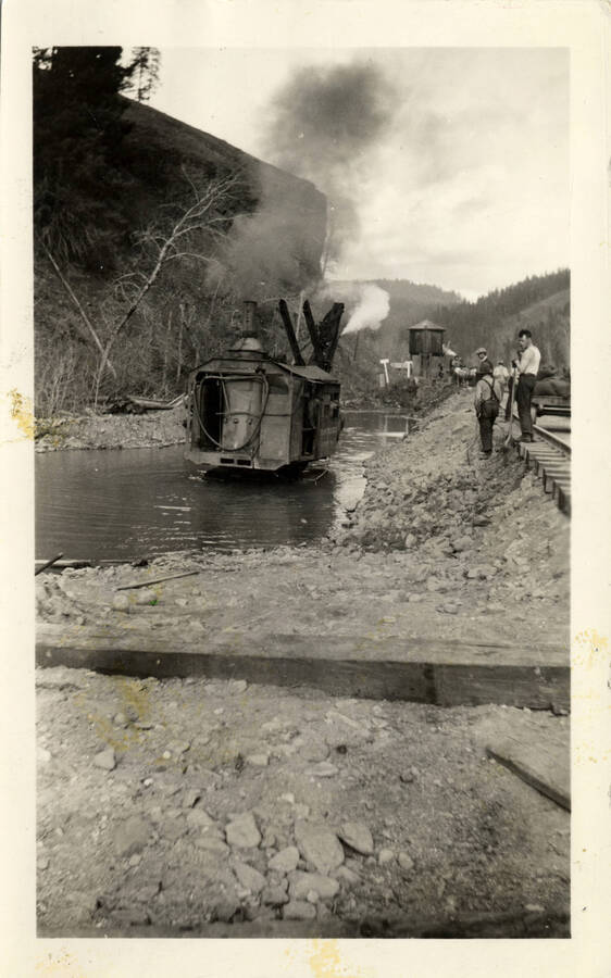 A black and white photograph of what is colloquially called a 'Logger Line' stuck in the water next to a pair of railroad tracks.