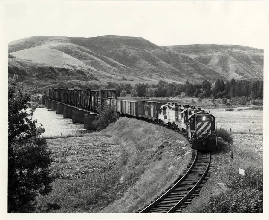 A photograph of train engine Extra 1703 East crossing the Clearwater River atop a metal bridge with ''commercials' ahead, log flats behind. The train is headed for Orofino. Mountains stretch across the background.