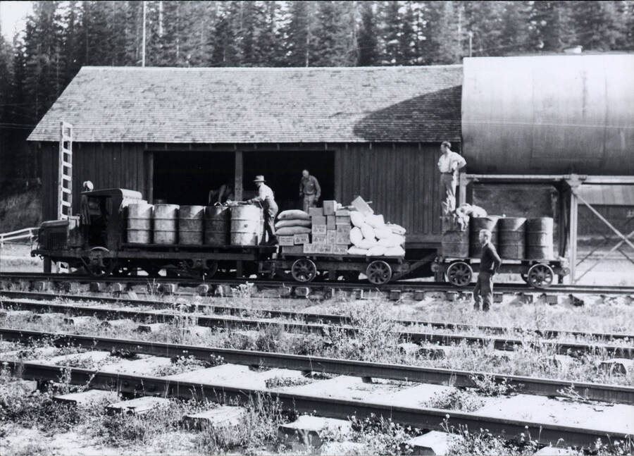 A photograph of a group of men loading large metal barrels, along with other supplies and materials, onto a small transport train. Located '3 and 1/2 miles South of Headquarters,' according to caption.
