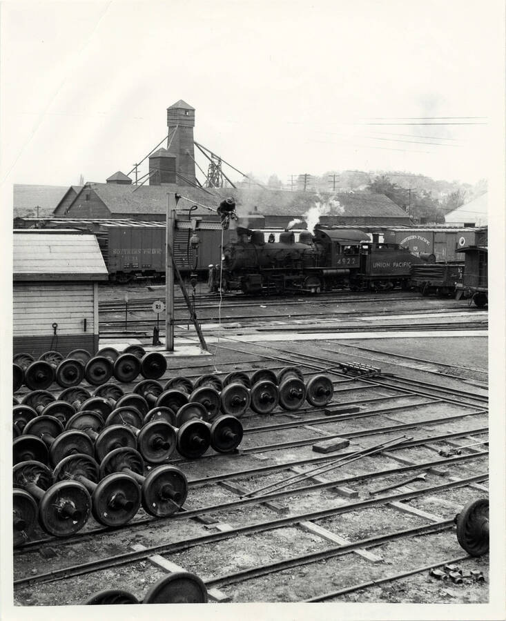 A photograph of the Lewiston Train Yard in Lewiston, ID. The photo shows train engine #4922 working the yard past the rip track area. An employee is repairing the wheel hoist in a manner that would have been frowned on by OSHA a generation later.