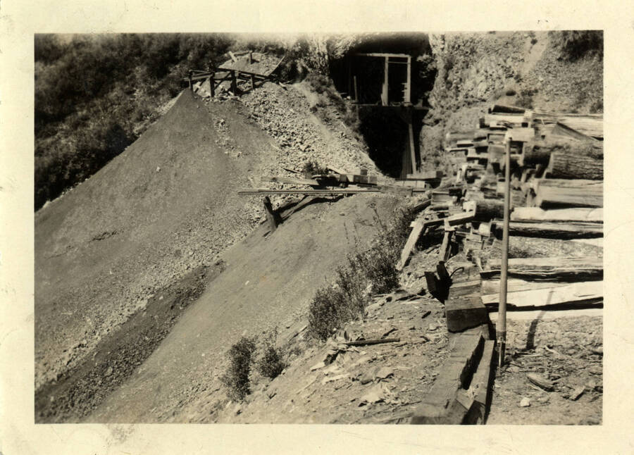 A photograph of the aftermath of weed burning in order to clear dirt for the railroad to be built. Also captured in the photo are the beginnings of a tunnel being created, and a stack of lumber waiting to be used to construct the railroad.