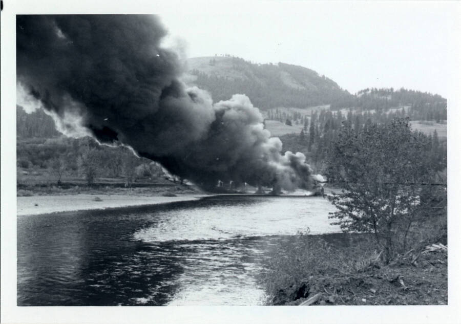 A photograph of Bridge 50.1 on the Camas Prairie Railroad next to the Kamiah Mine. Heavy black smoke billows from a fire on the railroad bridge.